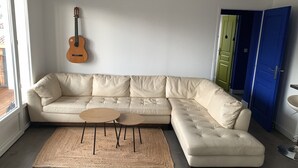 Ein Wohnzimmer mit einem beigefarbenen Ecksofa, einem kleinen runden Tisch, einer Gitarre an der Wand und einer blauen Tür.