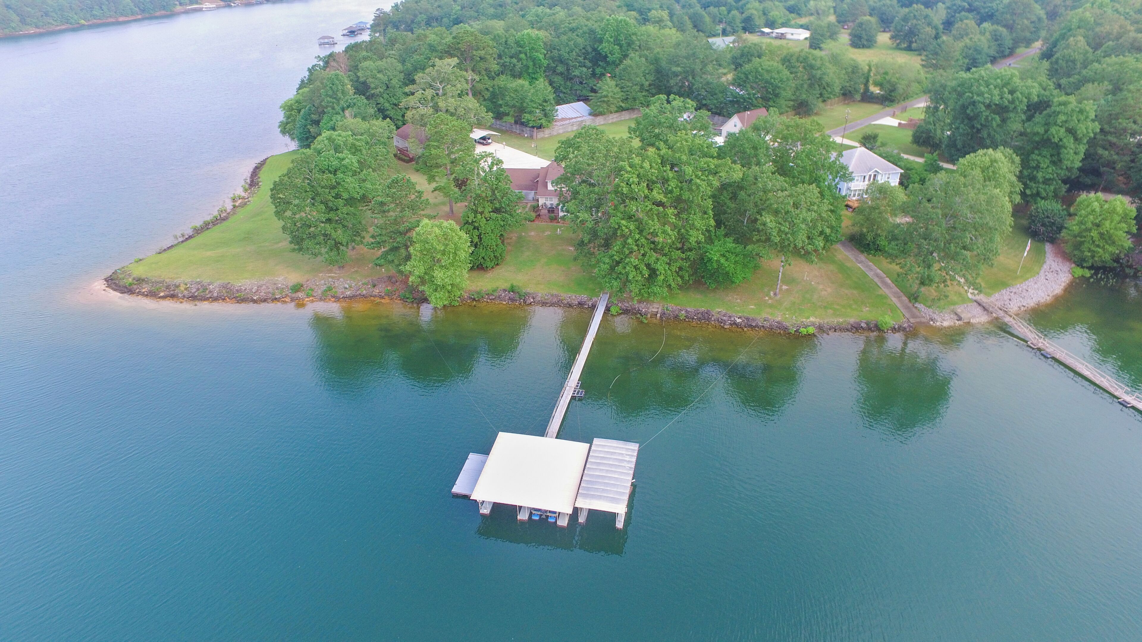 An aerial view of a lakeside property with a dock extending into the water, surrounded by lush greenery and residential buildings.