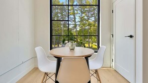 A room with a round table and two white chairs, a large window with a view of trees, and a white door. - 1BR Condo I King Bed + Sofa I Near DT TC (Traverse City)