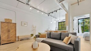 A modern living room with a grey sofa, a wooden table with a vase, and a kitchen in the background. - 1BR Condo I King Bed + Sofa I Near DT TC (Traverse City)