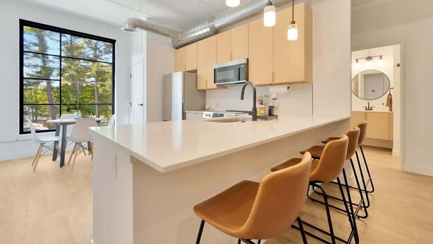A modern kitchen with a large island, bar stools, and a view of trees through large windows. - 1BR Condo I King Bed + Sofa I Near DT TC (Traverse City)