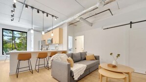 A modern living space with a grey sofa, wooden floor, and a kitchen area in the background. - 1BR Condo I King Bed + Sofa I Near DT TC (Traverse City)