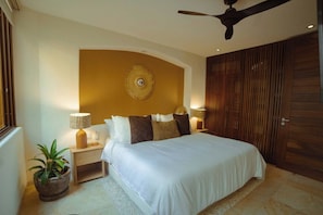 A bedroom with a bed, bedside lamps, a ceiling fan, a potted plant, and a wooden door.