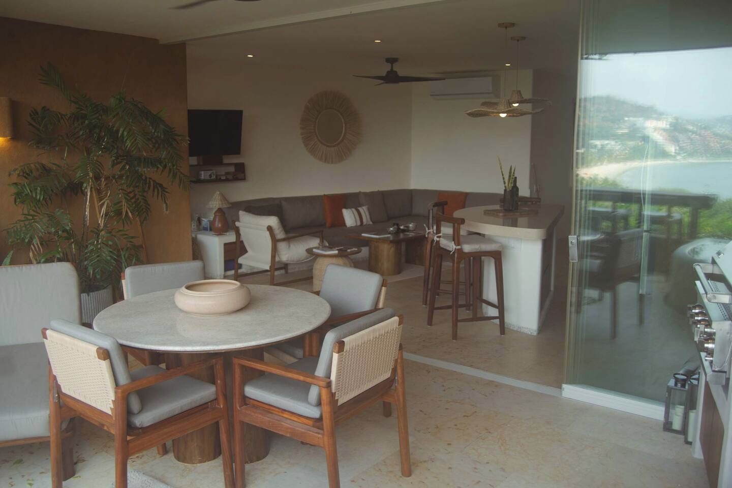 Una sala de estar moderna con una mesa de comedor redonda, asientos cómodos, una zona de cocina y un amplio ventanal corredero que da a un espacio exterior.