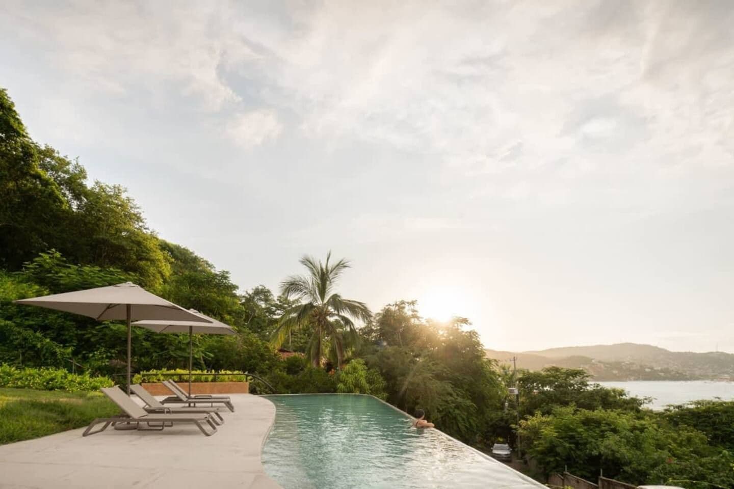 Une piscine dotée de chaises longues et d’un parasol, entourée d’une végétation luxuriante et offrant une vue sur la mer.