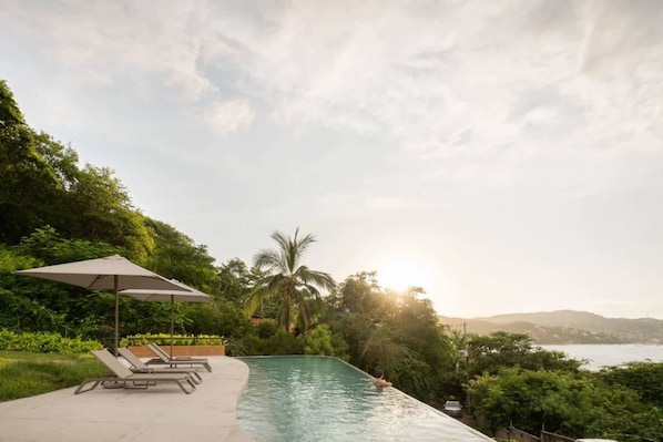 A swimming pool with lounge chairs and an umbrella, surrounded by lush greenery and a view of the sea.