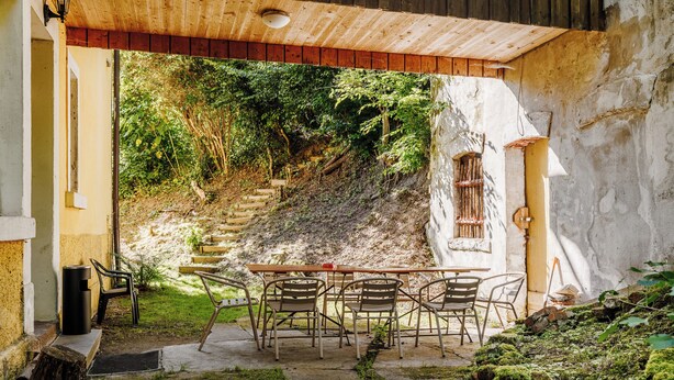 Um pátio externo com mesa e cadeiras, cercado por vegetação e uma escadaria de pedra.