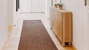 Um corredor com piso de madeira, tapete marrom, mesa consola de madeira com plantas em vasos e quadros nas paredes.