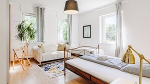 Quarto moderno com cama, duas áreas de estar, uma janela grande e um lustre.