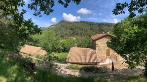 A rustic stone building with a tiled roof, surrounded by lush greenery and a mountainous backdrop.