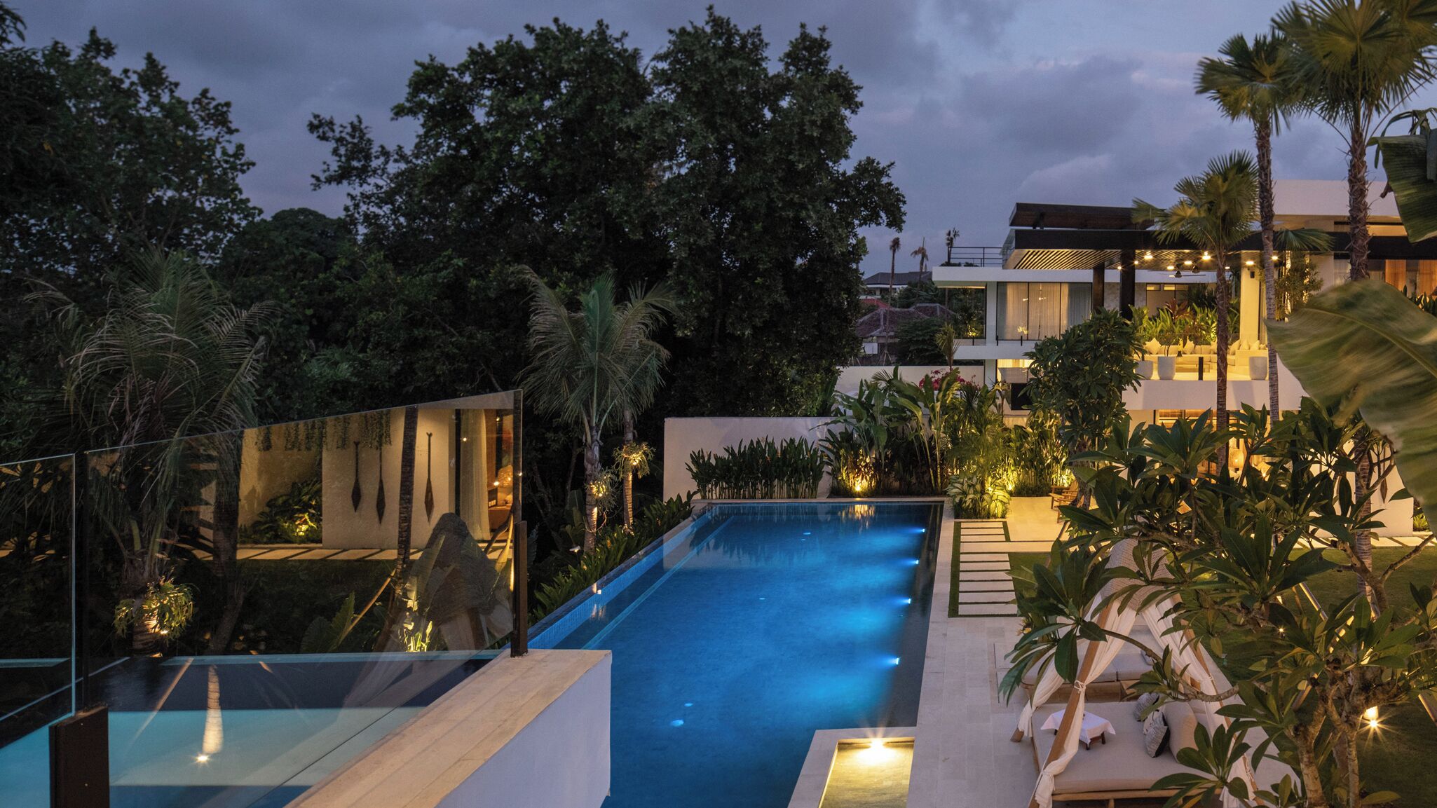 A modern outdoor swimming pool with a glass balustrade, surrounded by lush greenery and palm trees.
