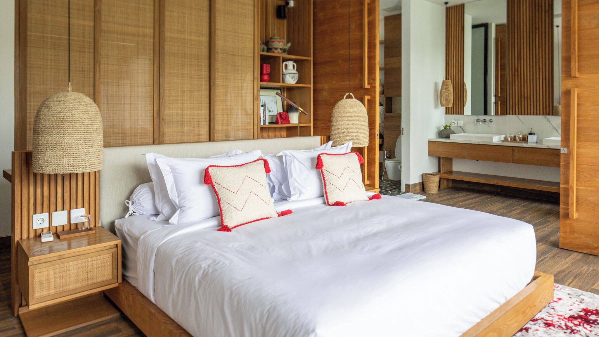 Un grand lit avec du linge de lit blanc et des oreillers à motifs rouges et blancs, une tête de lit en bois et une table de chevet surmontée d’une lampe en osier.