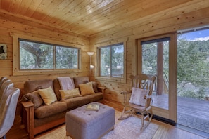 L’intérieur chaleureux d’un chalet avec un plafond en bois, un canapé, un fauteuil à bascule et de grandes fenêtres donnant sur la forêt.