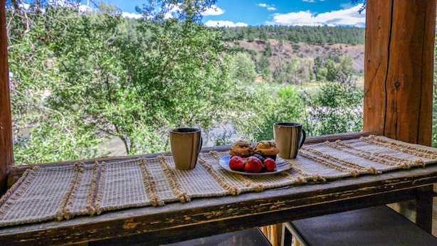 Una mesa de madera con un posavasos tejido, dos tazas, un plato con fresas y muffins, y una vista panorámica de árboles y colinas.