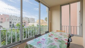 Un balcón con mesa y sillas, ventanas amplias y vistas a edificios y vegetación.