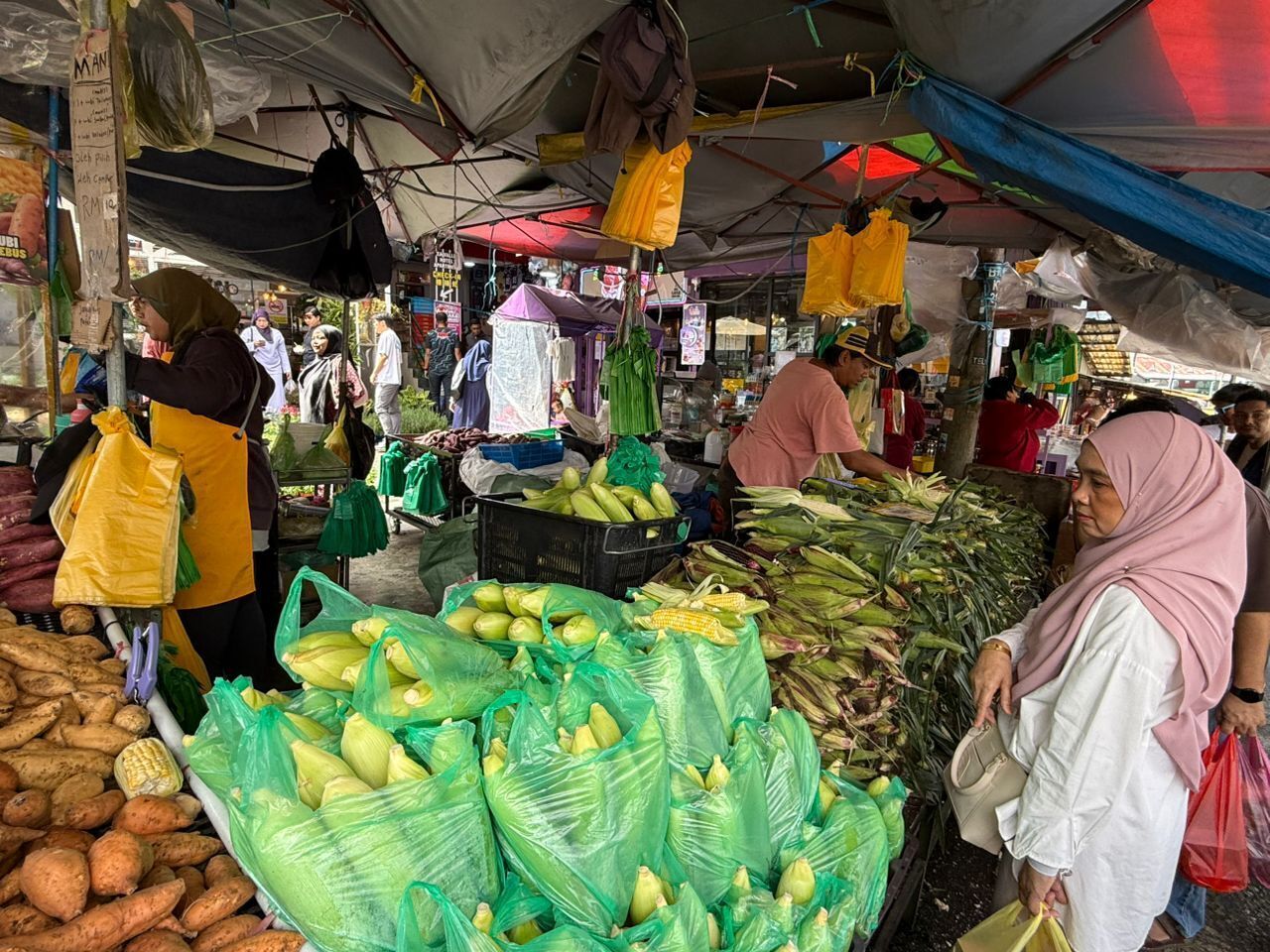 A market stall with various vegetables and fruits, including corn and potatoes, under a covered area.