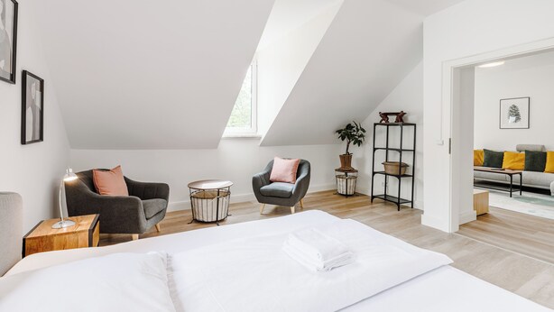 Une chambre moderne avec un grand lit, deux fauteuils, une petite table et une bibliothèque.