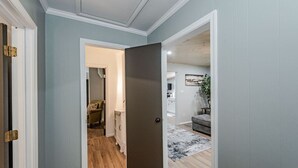 Un pasillo con suelo de madera, una puerta cerrada y vista a una sala de estar con un sofá gris y una planta.