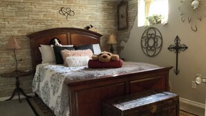 Een slaapkamer met een houten bed, een bankje aan het voeteneind, een bakstenen muur, een lamp en een decoratieve wandversiering.