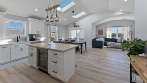 Een ruime keuken met een centraal eiland, een eetgedeelte met een tafel en stoelen, een woonkamer met een bank, en een groot raam dat zorgt voor natuurlijk licht.
