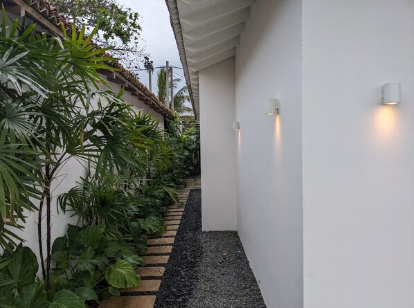 Un sendero estrecho bordeado por abundante vegetación y paredes blancas, que conduce a un edificio con tejado de tejas.