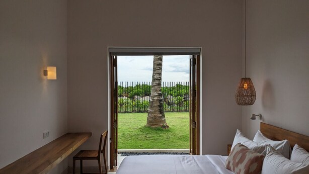 Un dormitorio con una cama grande, un banco de madera y vistas a un área verde con un árbol y una valla.