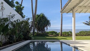 Una piscina con reflejo cristalino, rodeada de exuberante vegetación y palmeras, con un edificio blanco y una pérgola al fondo.