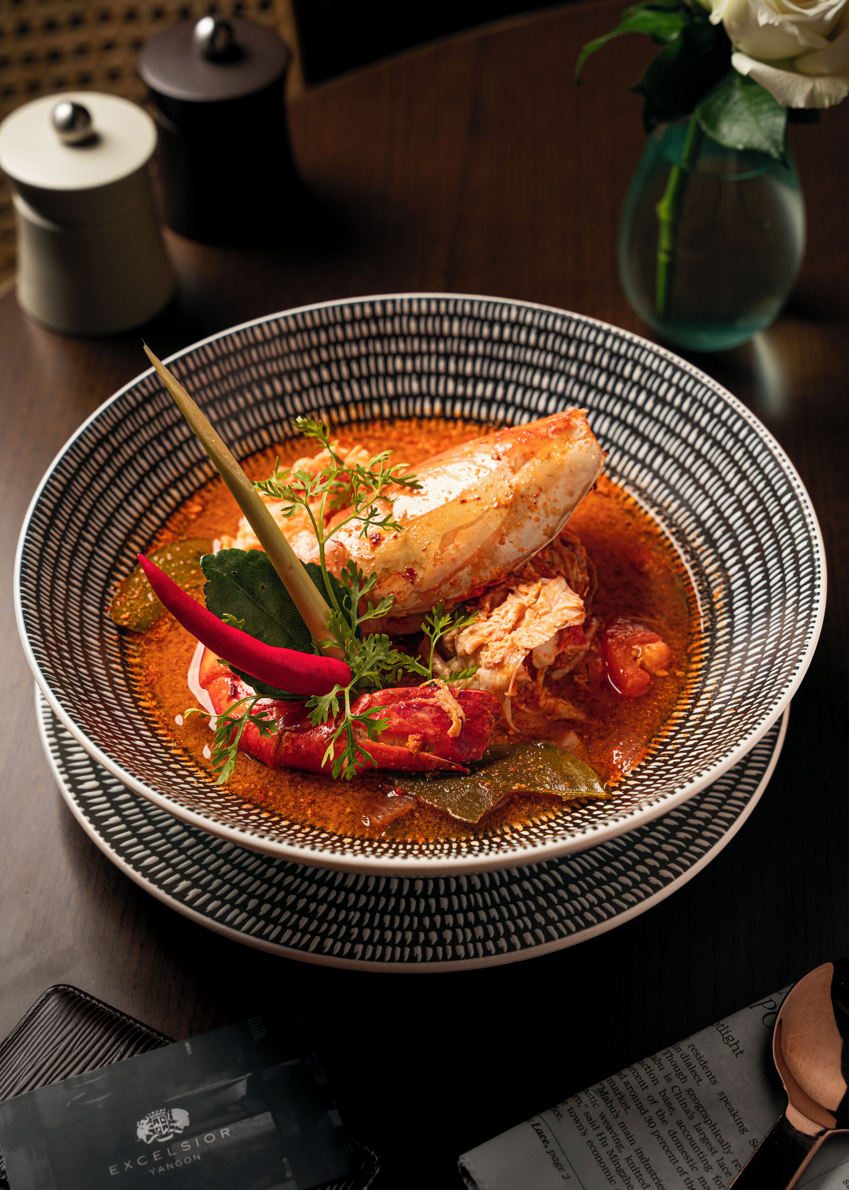 A bowl of food with a fish, vegetables, and a red chili.