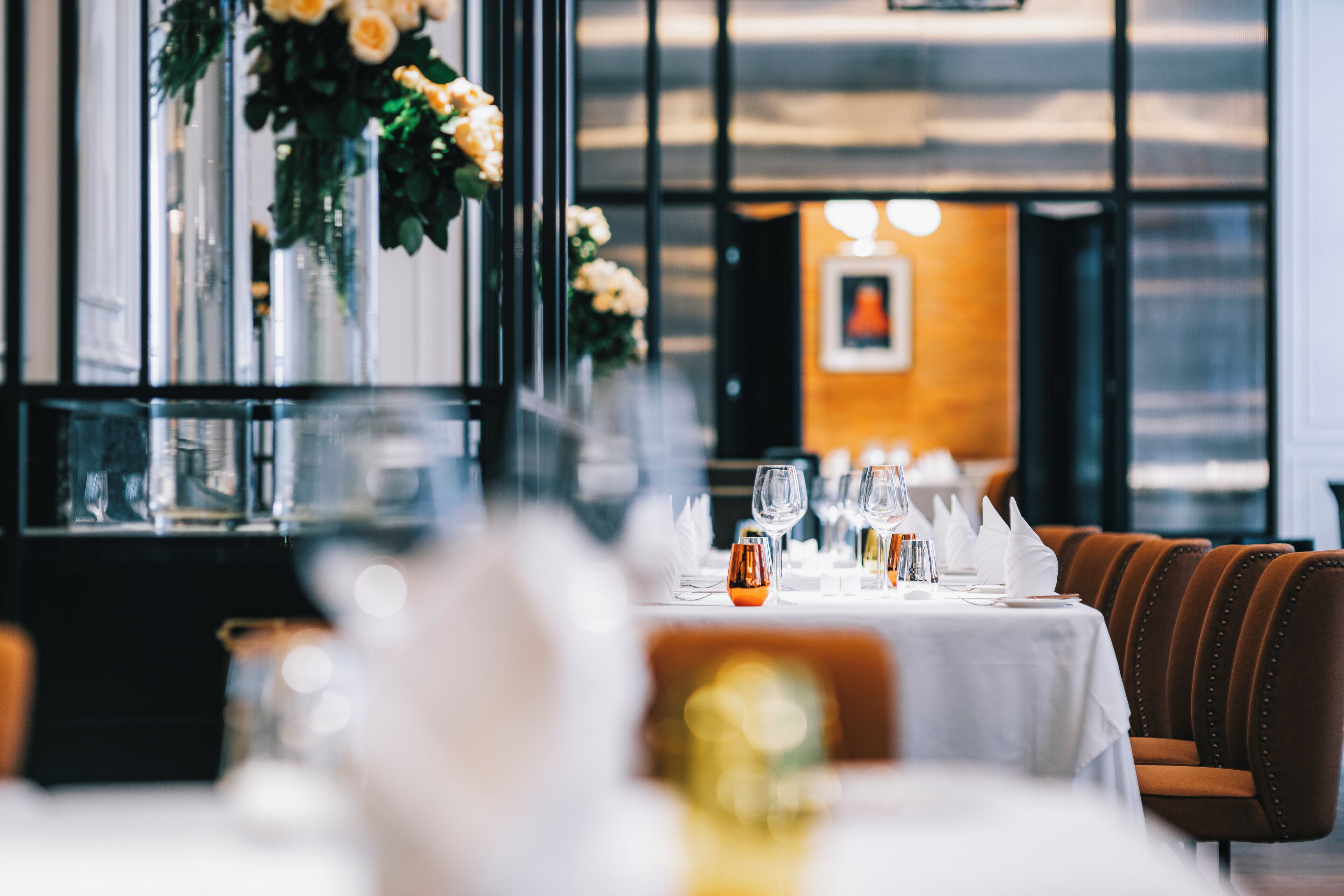 A restaurant with a long table set for dining, a vase of flowers, and a framed picture on the wall.