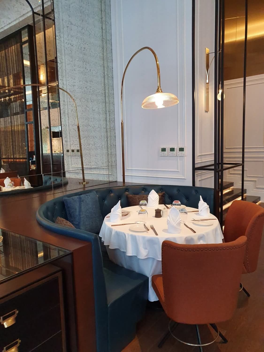 Een eetgedeelte met een ronde tafel gedekt voor twee personen, een blauwe gewatteerde bank en een hoge vloerlamp.