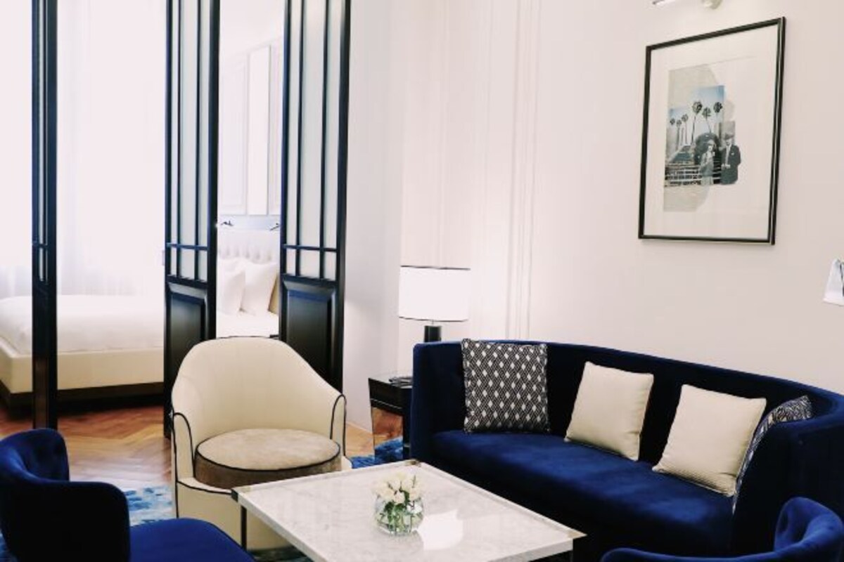 A modern hotel room with a bed, a blue sofa, a white armchair, a coffee table with a vase, and a framed picture on the wall.