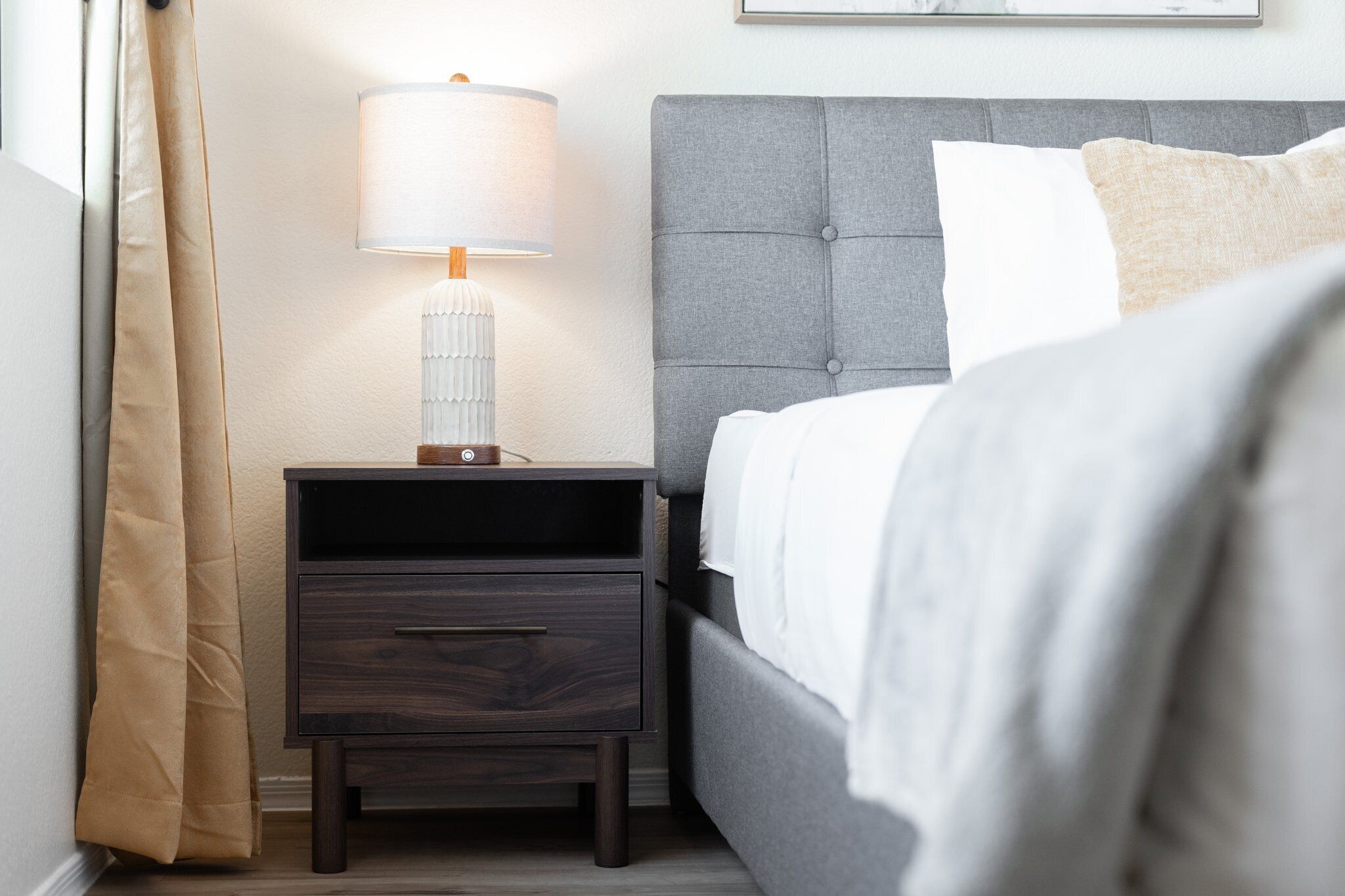 A neatly made bed with a grey headboard, a bedside table with a lamp, and a brown curtain.