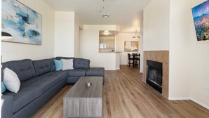 Un salon moderne avec un canapé d’angle, une table basse, une cheminée et un tableau représentant un paysage urbain.