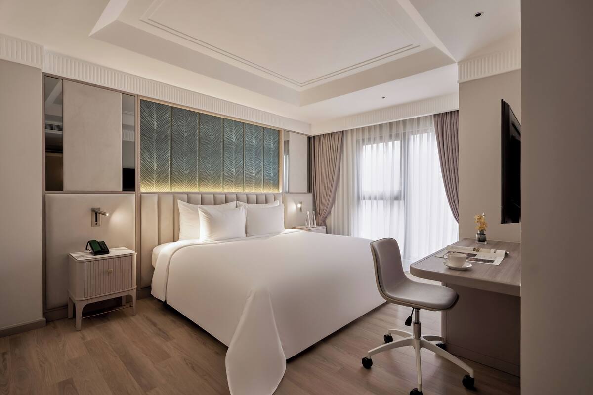 A modern hotel room with a large bed, a desk with a chair, a nightstand, and a large window with curtains.