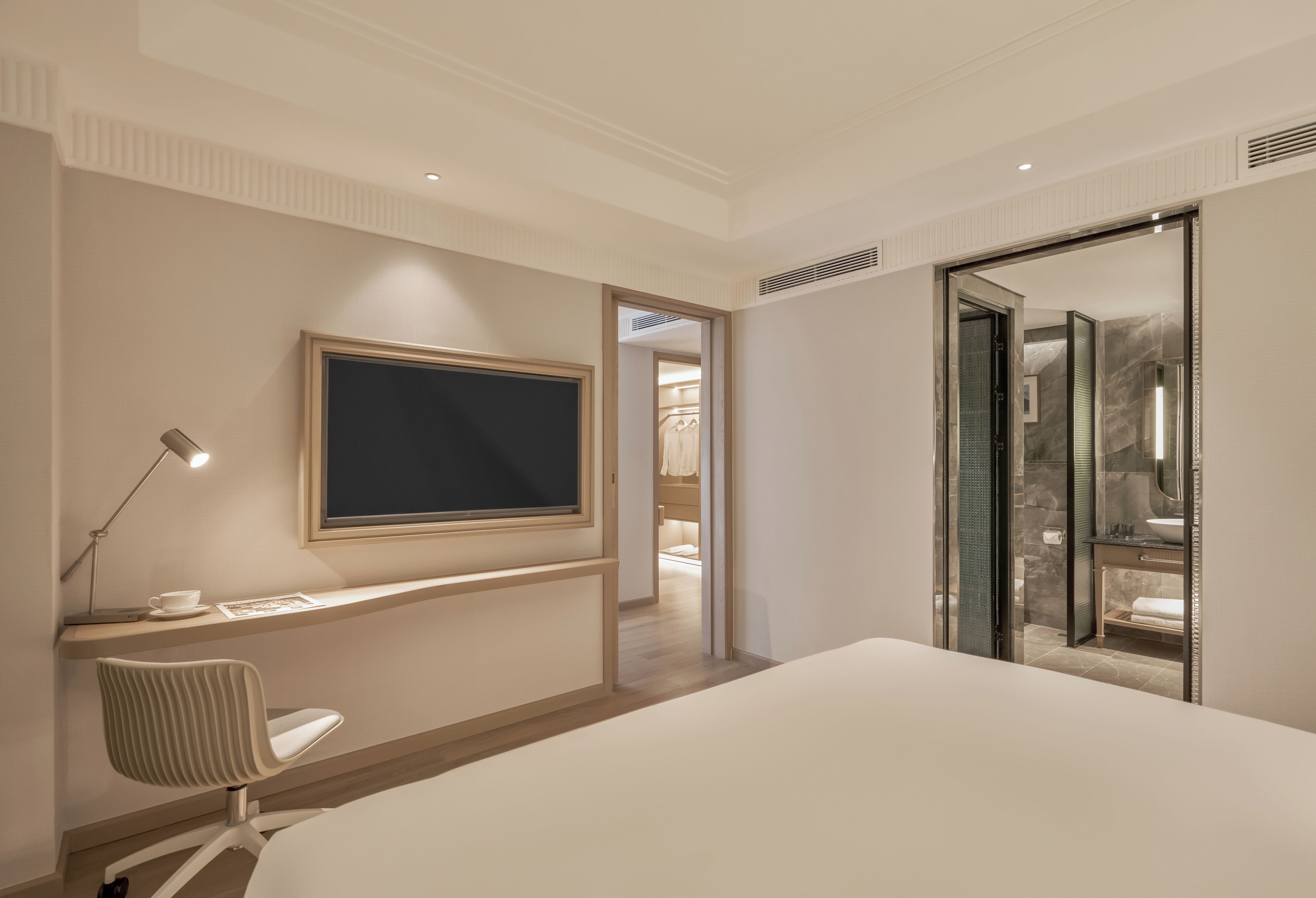 A modern hotel room with a bed, a desk with a chair, a flat-screen TV, and a bathroom visible through an open door.