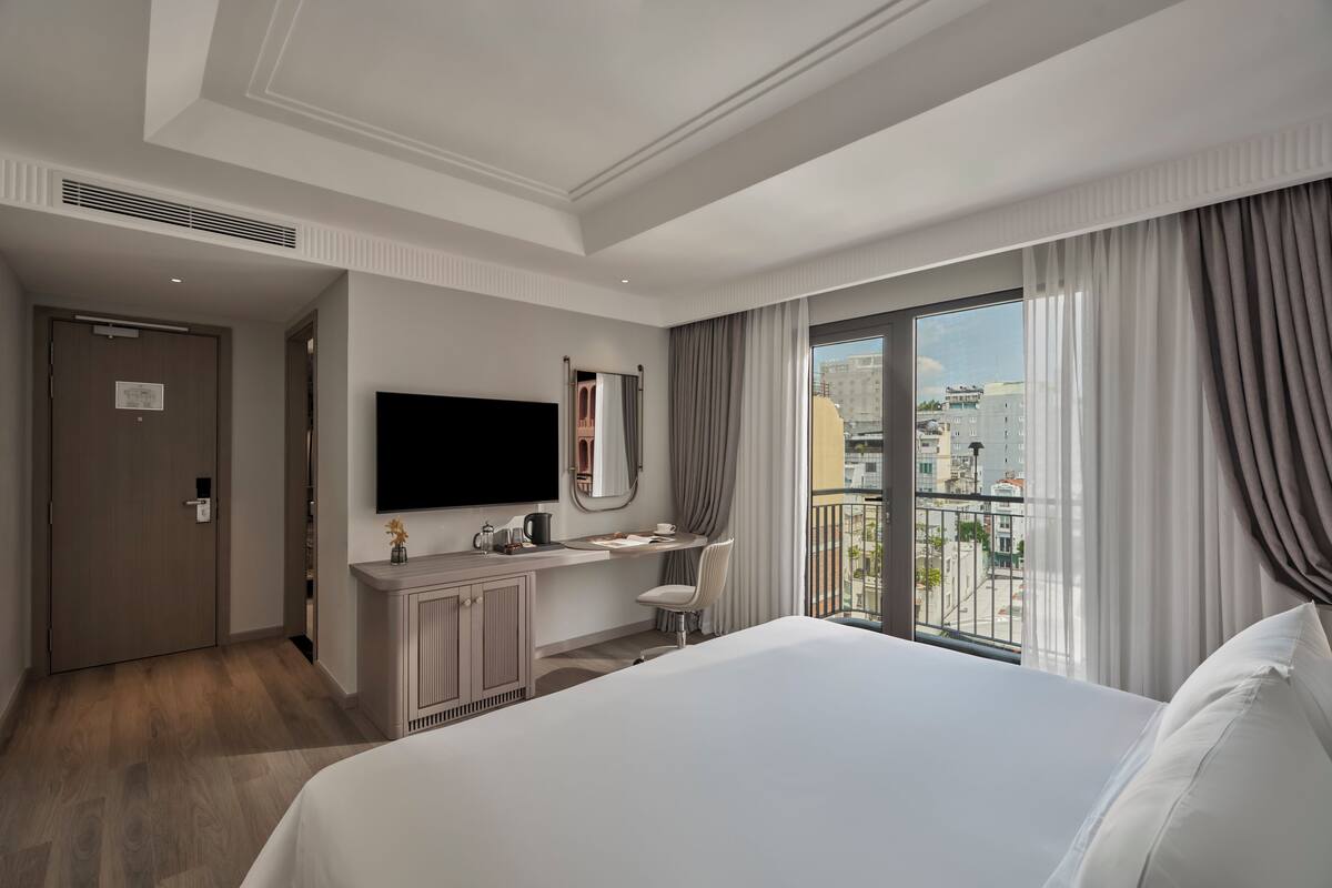 A hotel room with a large bed, a desk with a chair, a flat-screen TV, a window with curtains, and a view of the city.