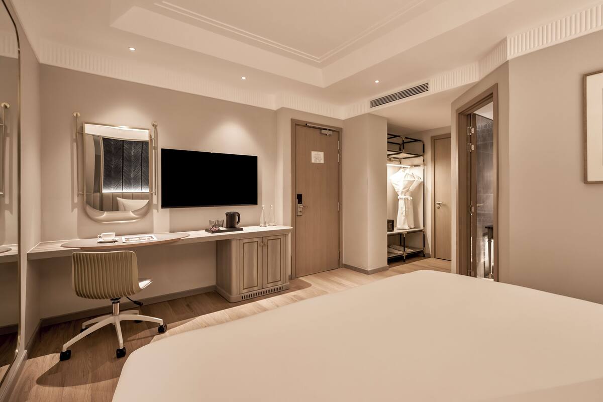 A modern hotel bedroom with a large bed, a desk with a flat-screen TV, a chair, and a walk-in closet.