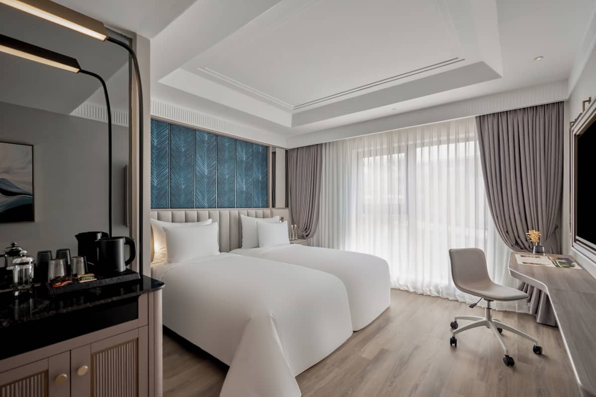 A modern hotel room with two beds, a desk, a chair, and a large window with curtains.