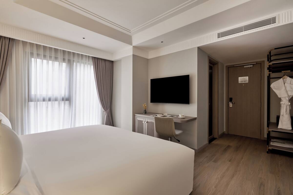 A hotel room with a large bed, a desk with a flat-screen TV, a wardrobe, and a door leading to another room.