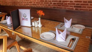 A restaurant setting with a brick wall, a menu stand, and a table set with plates, napkins, and a vase with flowers. - Hotel Garuda Bhaktapur (Bhaktapur)