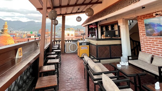 An outdoor seating area with wooden tables and benches, a bar with a Coca-Cola sign, and a view of a cityscape with mountains in the background. - Hotel Garuda Bhaktapur (Bhaktapur)