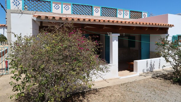 A white building with a blue and beige roof, a covered porch, and a small garden area with pink flowers. - Appartamento in Casa Paiesa, San Fernando Dista in 5 Minuti (Formentera Islas Baleares)