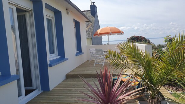 A wooden deck with a white and blue building, a patio umbrella, and potted plants. - The old Caon grocery store (Telgruc-sur-Mer)