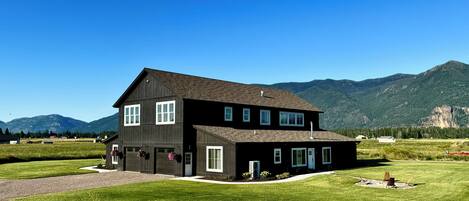 Une maison noire à deux étages avec un garage, entourée d’une pelouse bien entretenue et avec des montagnes en arrière-plan.