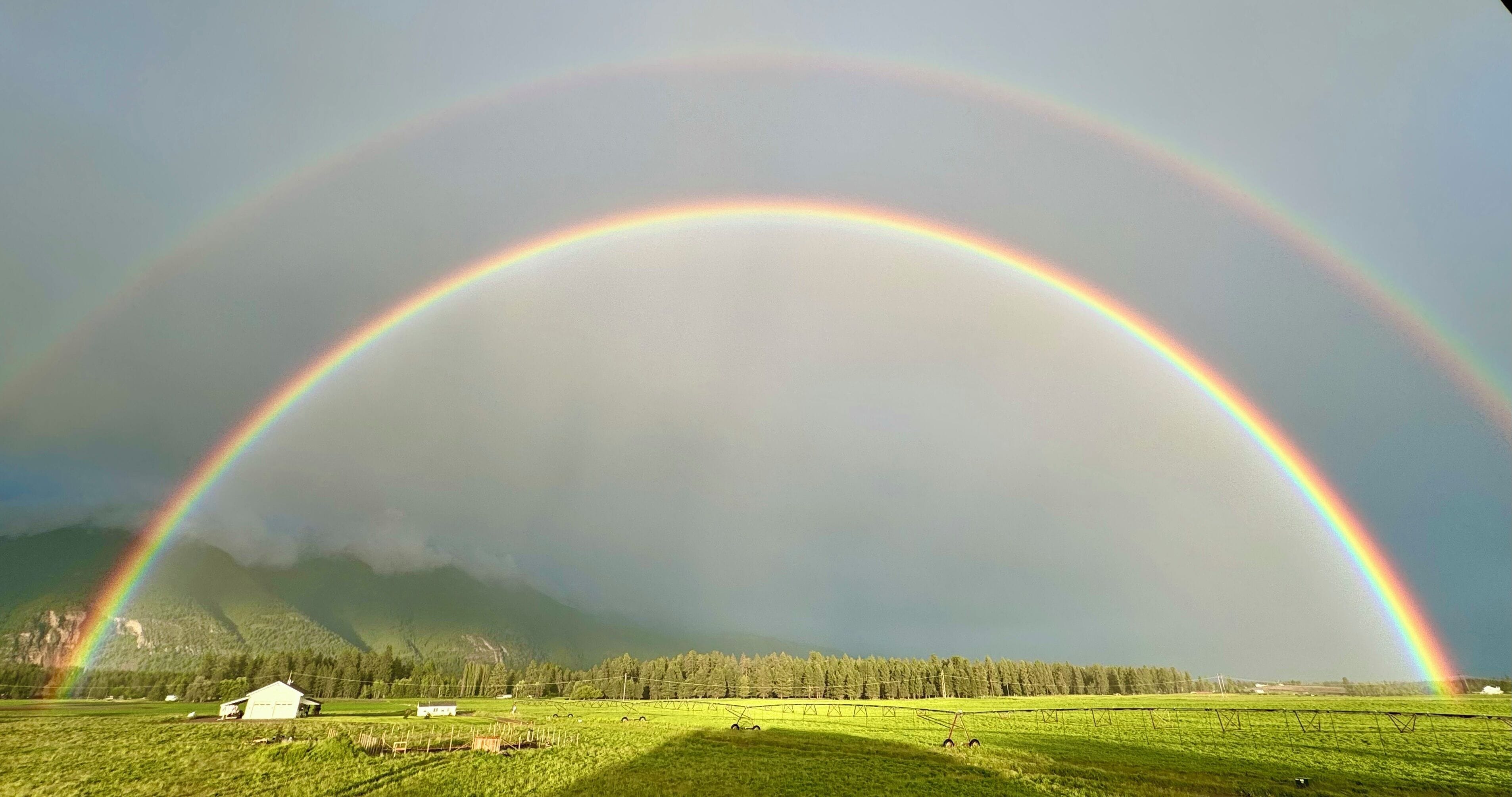 Un paysage rural avec un arc-en-ciel qui s’étend au-dessus d’un champ et un petit bâtiment au premier plan.