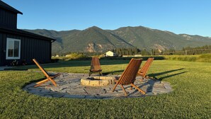 Un foyer en pierre, entouré de chaises en bois, se trouve dans une cour verdoyante, avec une montagne en toile de fond.