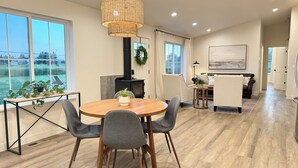 Un salon moderne avec une table ronde en bois, deux fauteuils gris, un poêle à bois noir et une grande fenêtre donnant sur un paysage verdoyant.