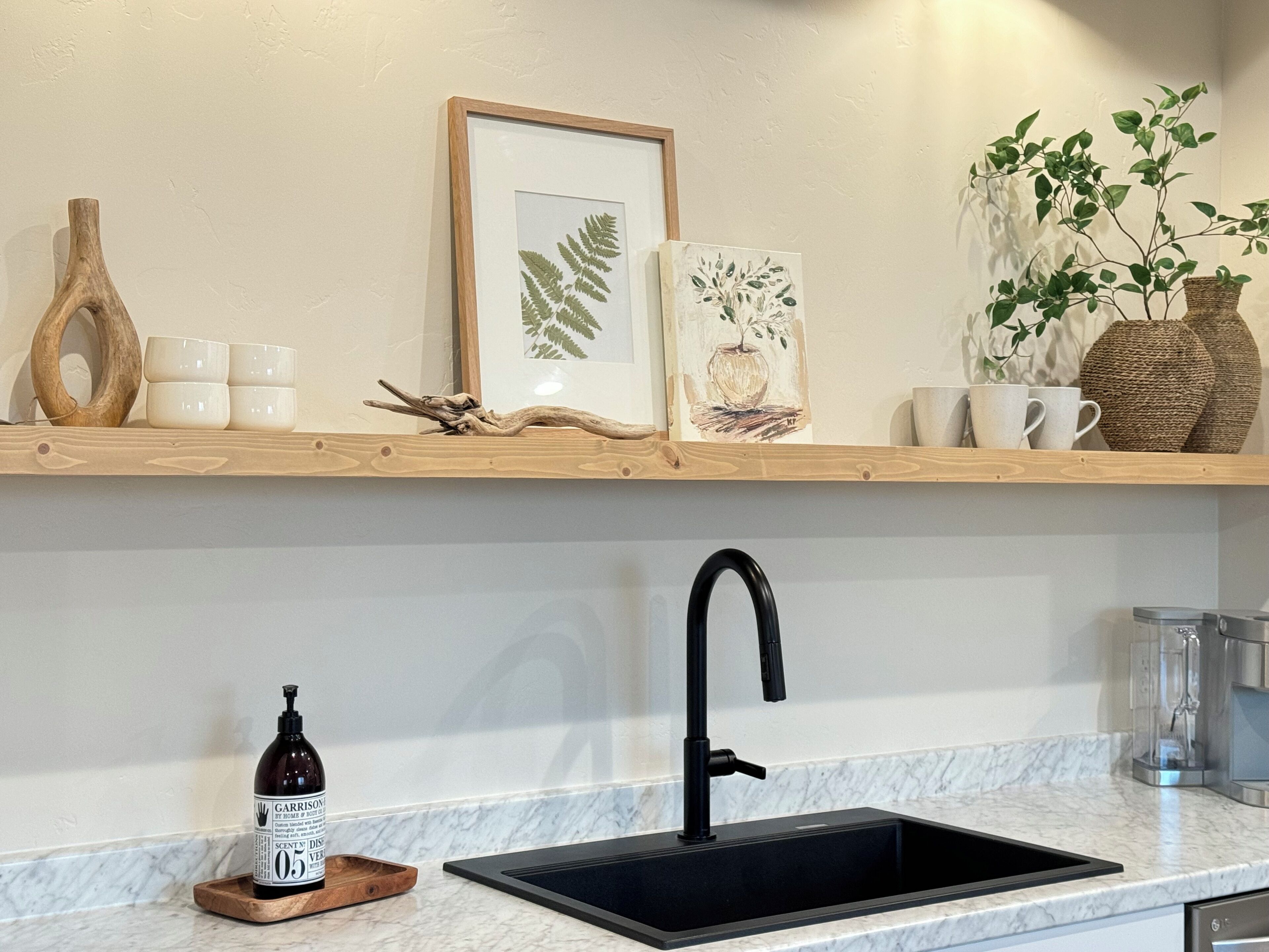 Una cocina con un lavamanos negro, encimera de mármol y estanterías de madera con objetos decorativos y un cuadro enmarcado.