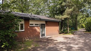 A small brick building with a door and windows, surrounded by trees and a gravel path. - Detached bungalow for 6 persons (Putten)