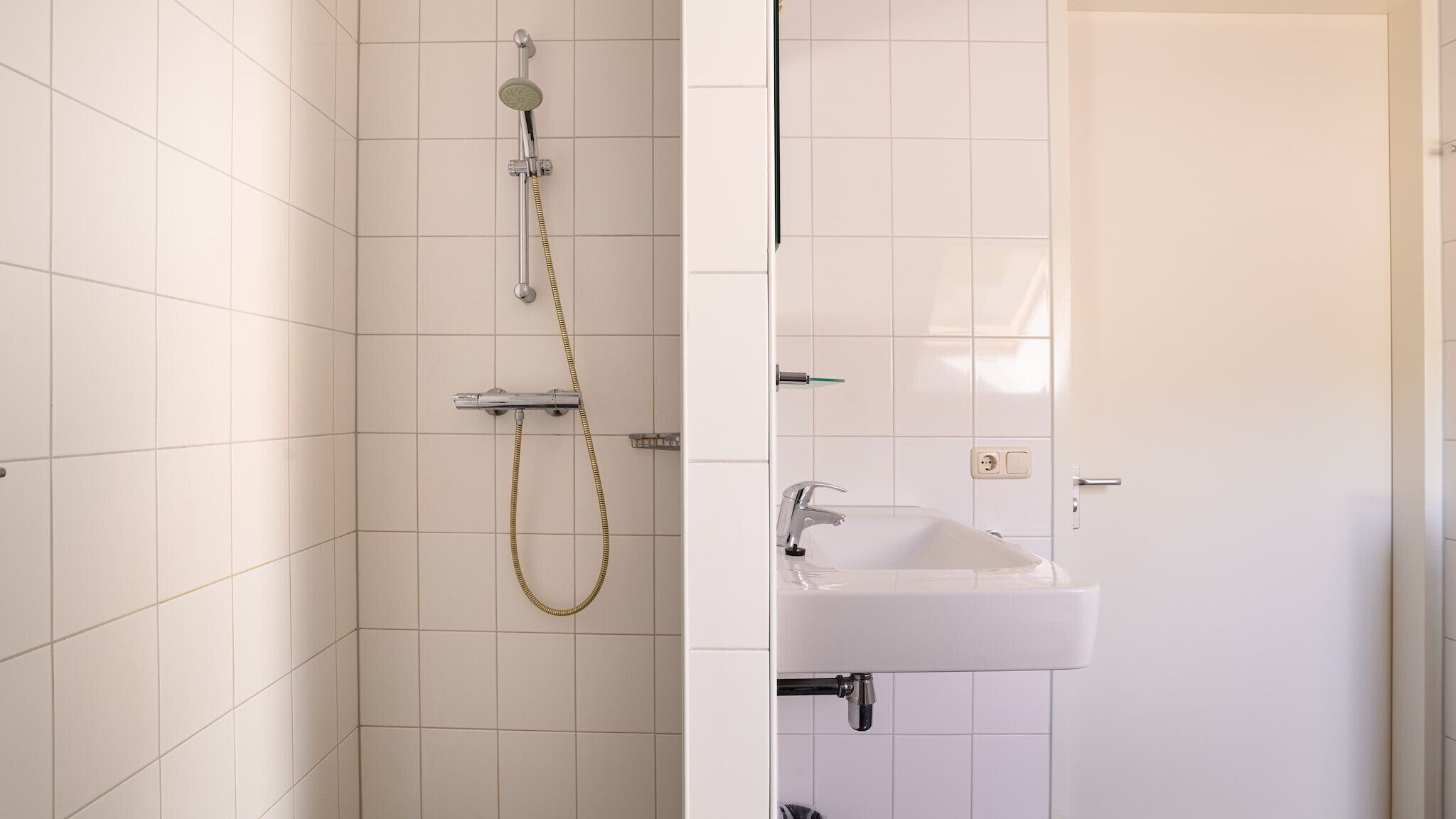 A bathroom with white tiled walls, a shower with a handheld showerhead, a sink with a faucet, and a door with a handle.